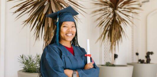 woman in academic gown holding a rolled certificate while smiling at the camera